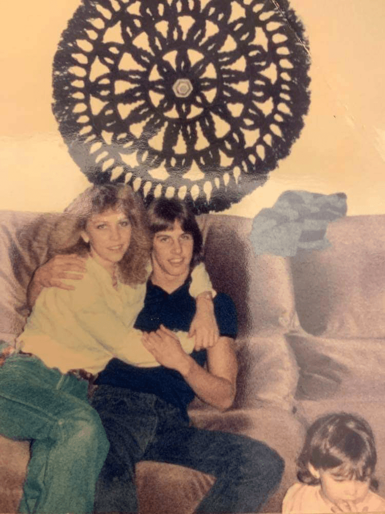 Vintage photo of Dani and Dennis seated on a sofa with a large medalion wall hanging behind and toddler seated on the floor in the foreground
