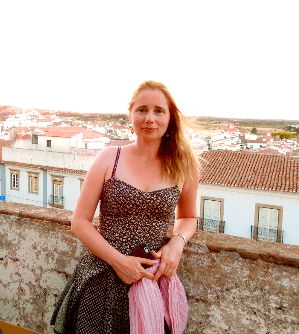 Elena in a sundress leaning on blacony wall with view of a city with terra cotta roof tiles below