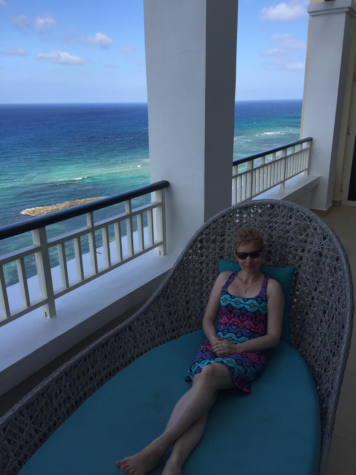 Elena seated on a large cane back chaise lounge on an oceanside patio. 