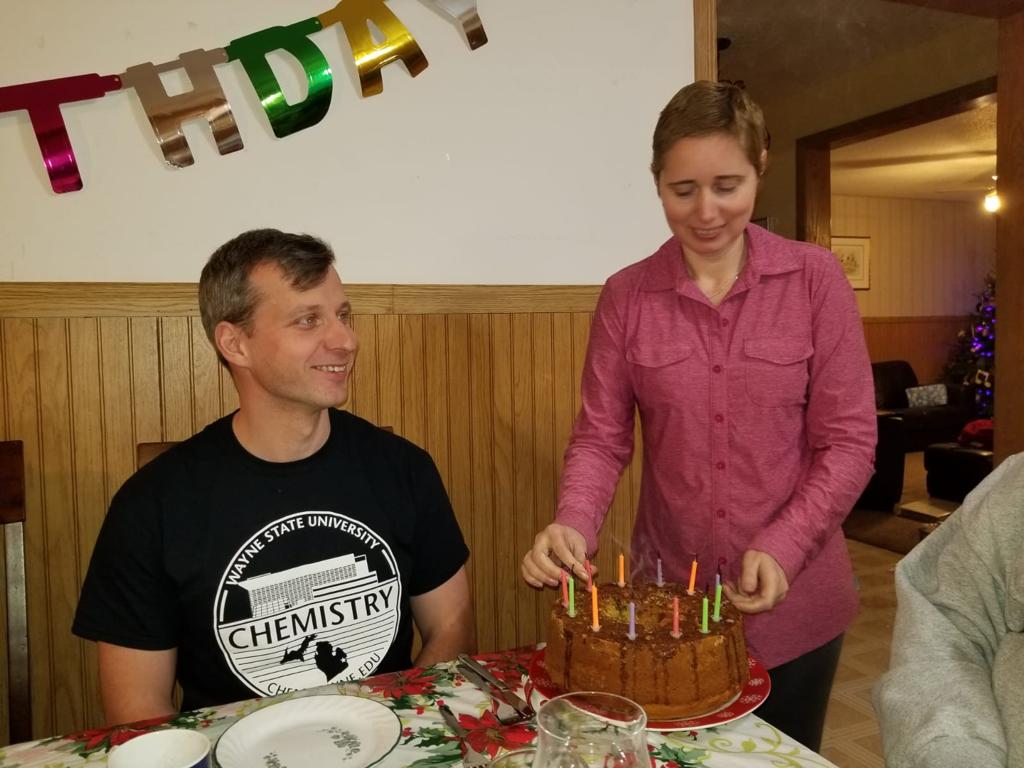 Pavel seated wearing Wayne State University Chemistry T-shirt and Elena stnding next to him removing birthday candles from a chocolate iced cake.