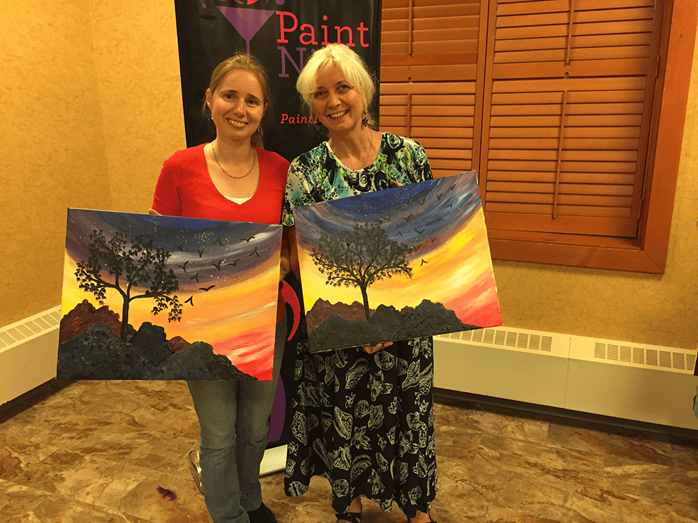 Elena and her mother, each holding a painting of a tree with birds flying in the sunset 